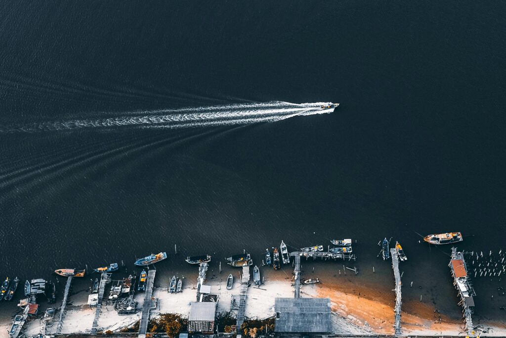 Drone shot capturing a motorboat speeding on dark water near a harbor with docked boats and sandy beach.